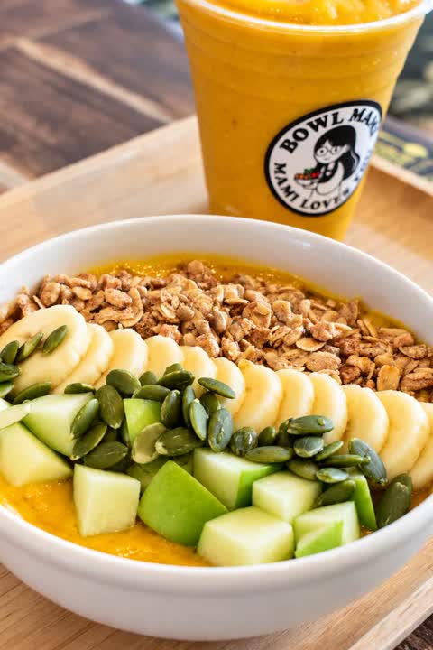 Yellow smoothie bowl topped with banana, green fruit, granola and pumpkin seeds, with a yellow smoothie
